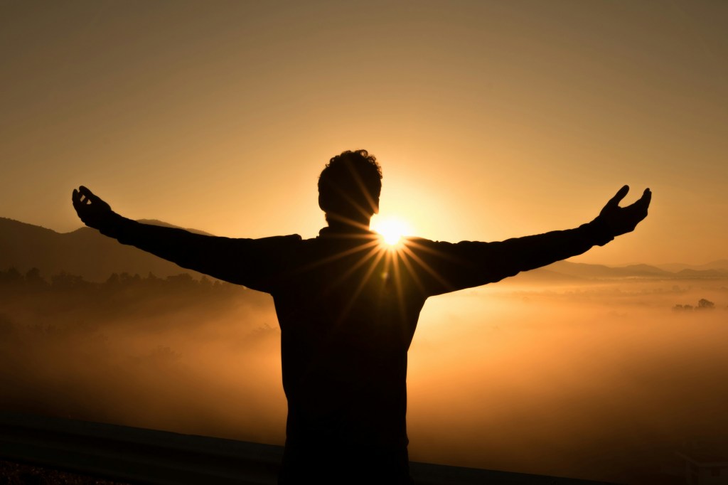 Person looking at rising sun with arms spread wide open.